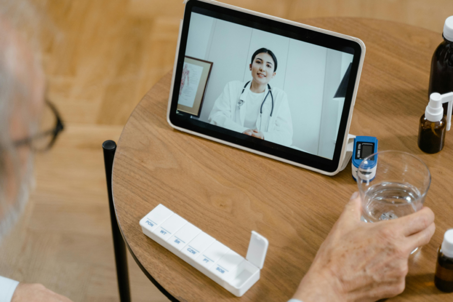 Elderly man on telehealth call with doctor, holding medication organizer and water.