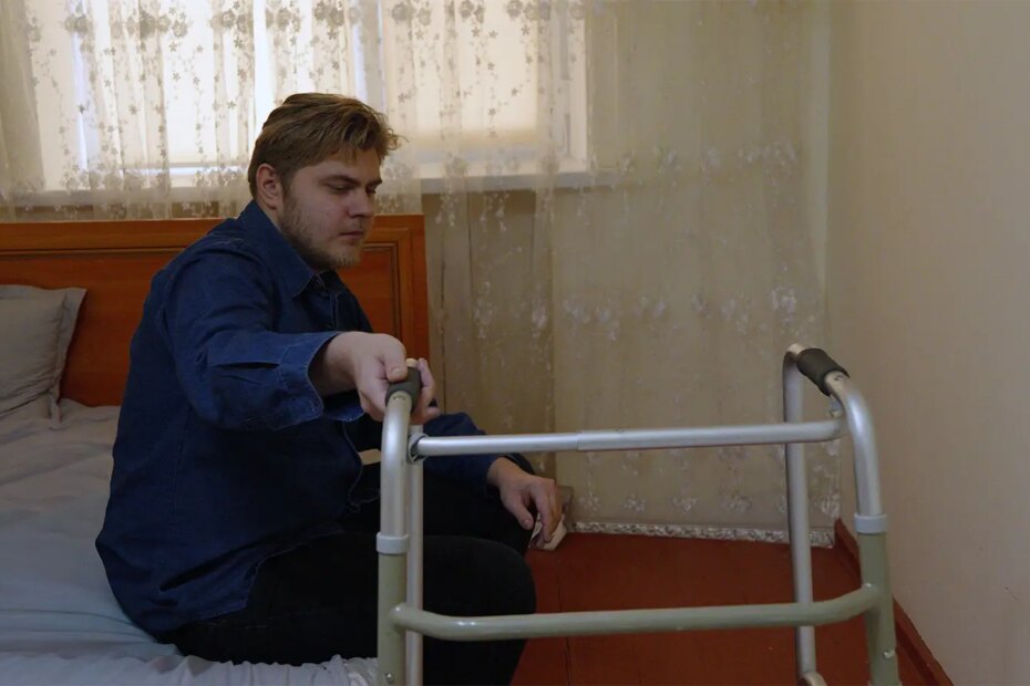 A photo of a man sitting on the edge of a bed and holding a walker.