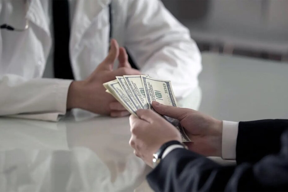 A photo of a man in a business suit holding a fan of $100 bills in front of a male physician.