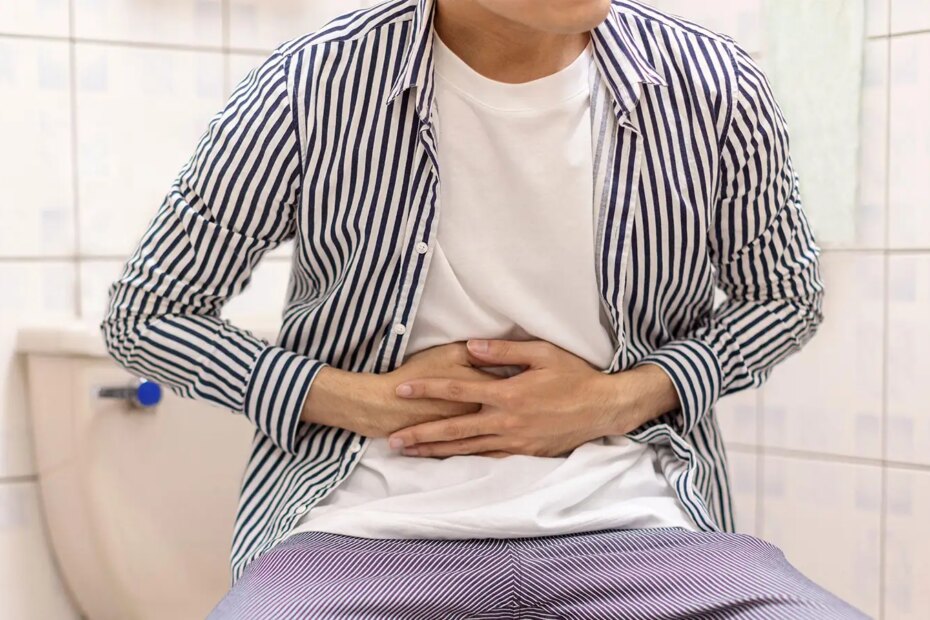 A photo of a fully clothed man sitting on a toilet and holding his stomach.