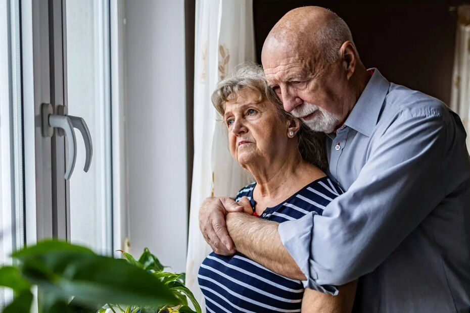 A photo of a senior man hugging his wife in front of a window in their home.