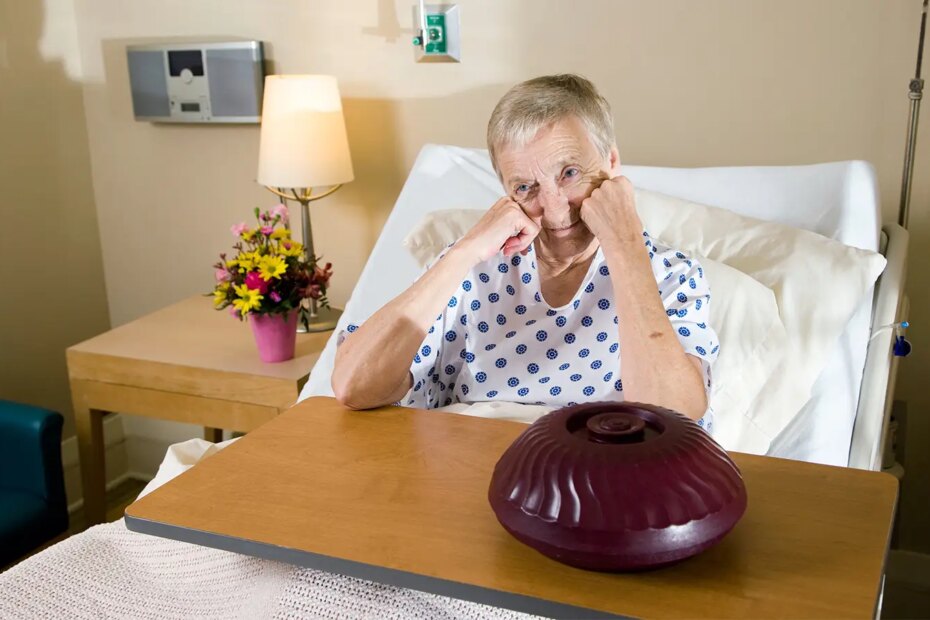 A photo of a senior woman in a hospital bed sitting with her arms resting on the tray table above a food container.