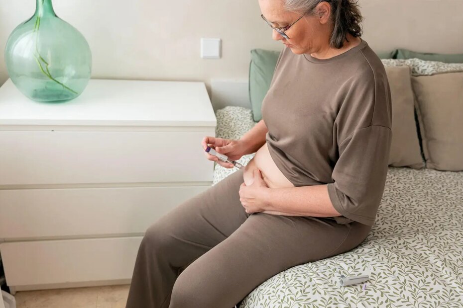 A photo of a mature woman sitting on the edge of a bed and self injecting in her abdomen.
